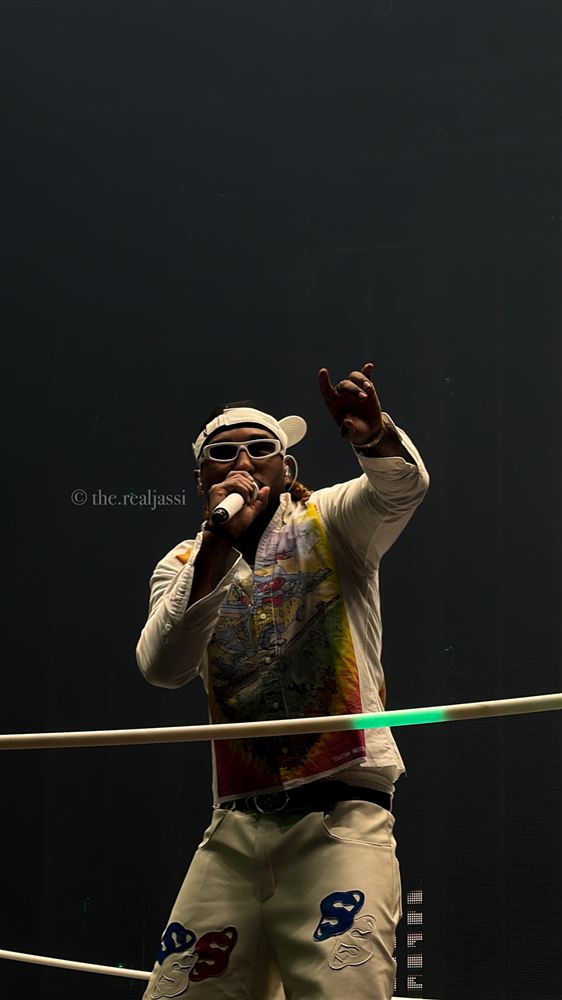 Reezy performing in a printed light jacket, cap and white sunglasses behind the ring ropes.