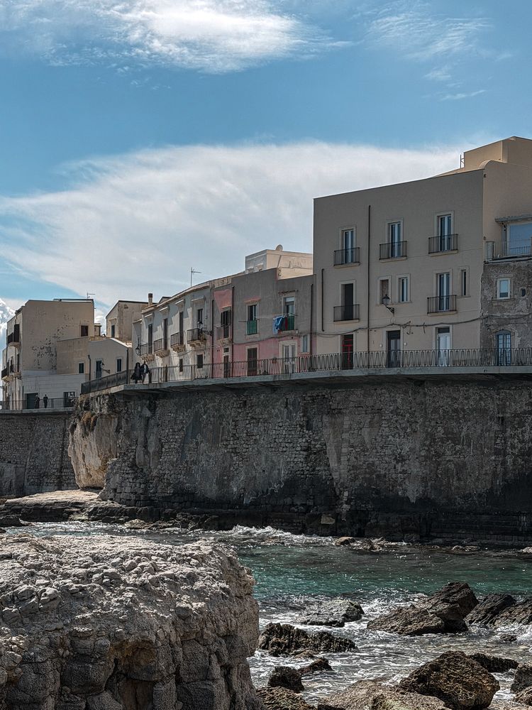 Seafront houses and balconies above a stone seawall, with rocks and turquoise water in Ortigia, Syracuse.